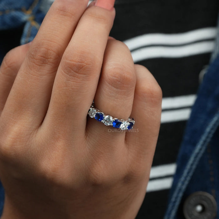 Blue Sapphire Cushion And Round Cut Diamond Eternity Wedding Band worn on a hand, highlighting the alternating blue sapphires and round diamonds, with a background showing a striped shirt and denim jacket.