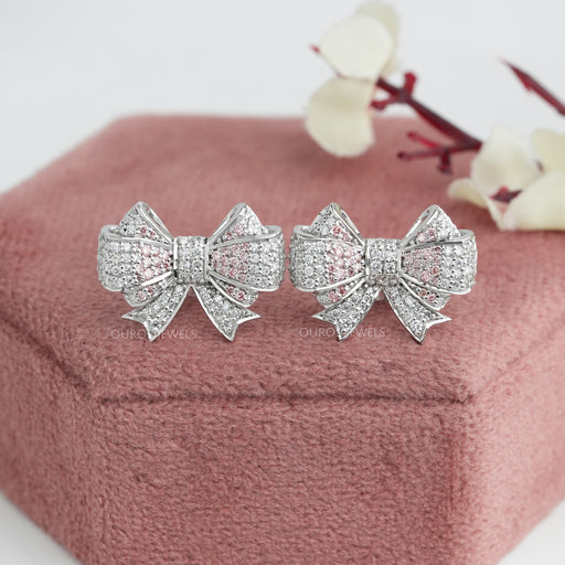 Bow Shaped Round Stud Diamond Earrings displayed on pink velvet box with blurred floral background.