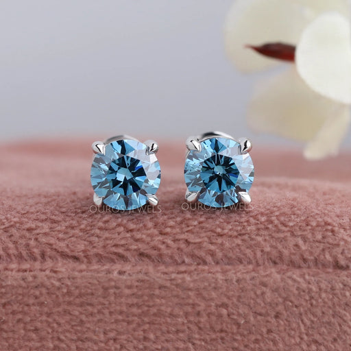 Blue Round Diamond Stud Earrings displayed on pink velvet box with blurred floral background.
